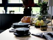 Schräg von vorne fotografierte, stilvoll gedeckte Tischszene mit schwarzen Keramikschalen und Tellern, Weinglas mit Weißwein, gerippten Trinkgläsern, Brotlaib und Birnen neben einer Glasvase mit Blumen auf hellem Holztisch.