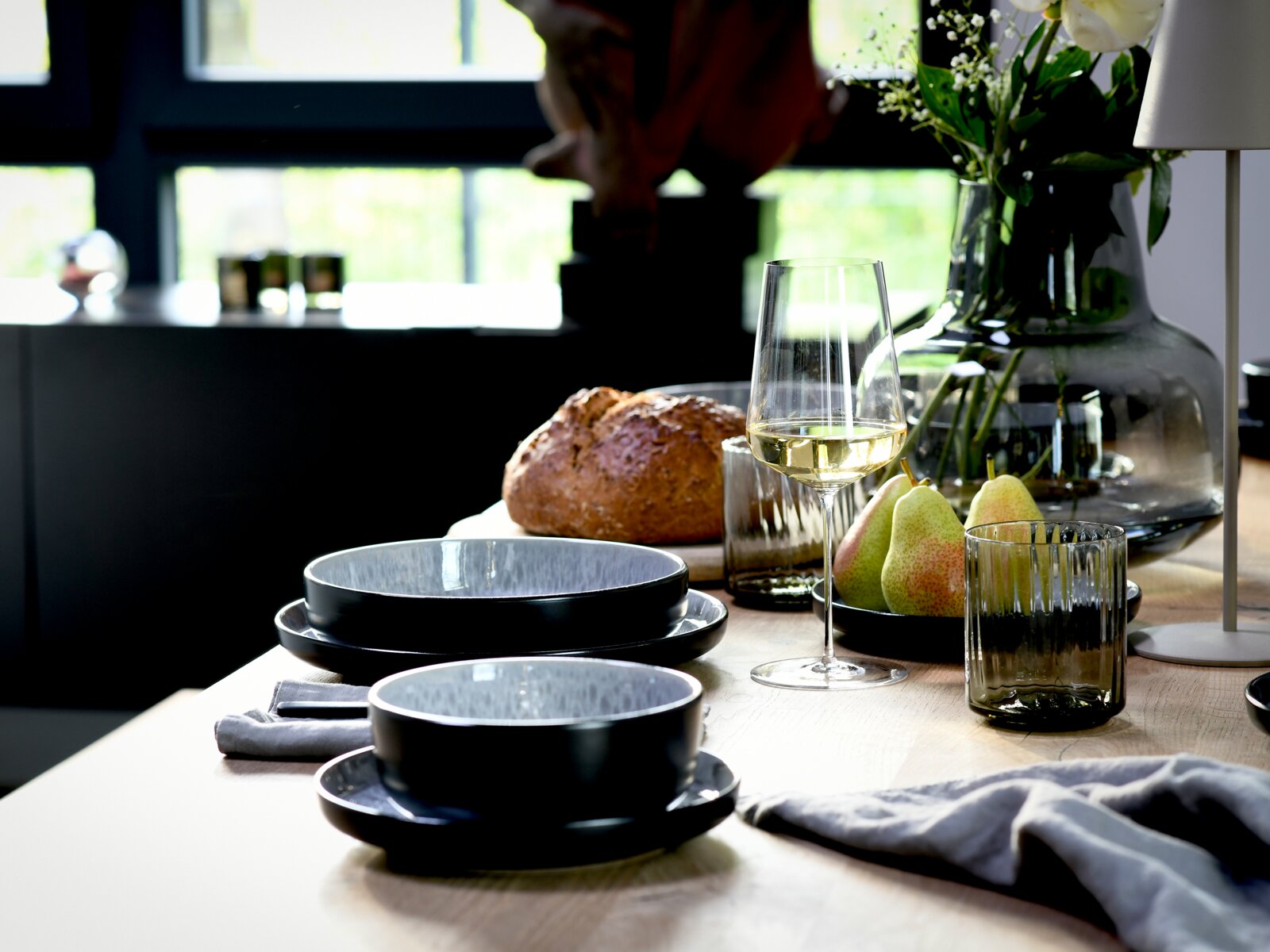 Schräg von vorne fotografierte, stilvoll gedeckte Tischszene mit schwarzen Keramikschalen und Tellern, Weinglas mit Weißwein, gerippten Trinkgläsern, Brotlaib und Birnen neben einer Glasvase mit Blumen auf hellem Holztisch.