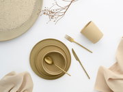 Minimalistisches beiges Keramik-Geschirrset mit Tellerstapel, Schale und Becher sowie goldfarbenem Besteck auf weißem Hintergrund, Draufsicht (Flatlay)