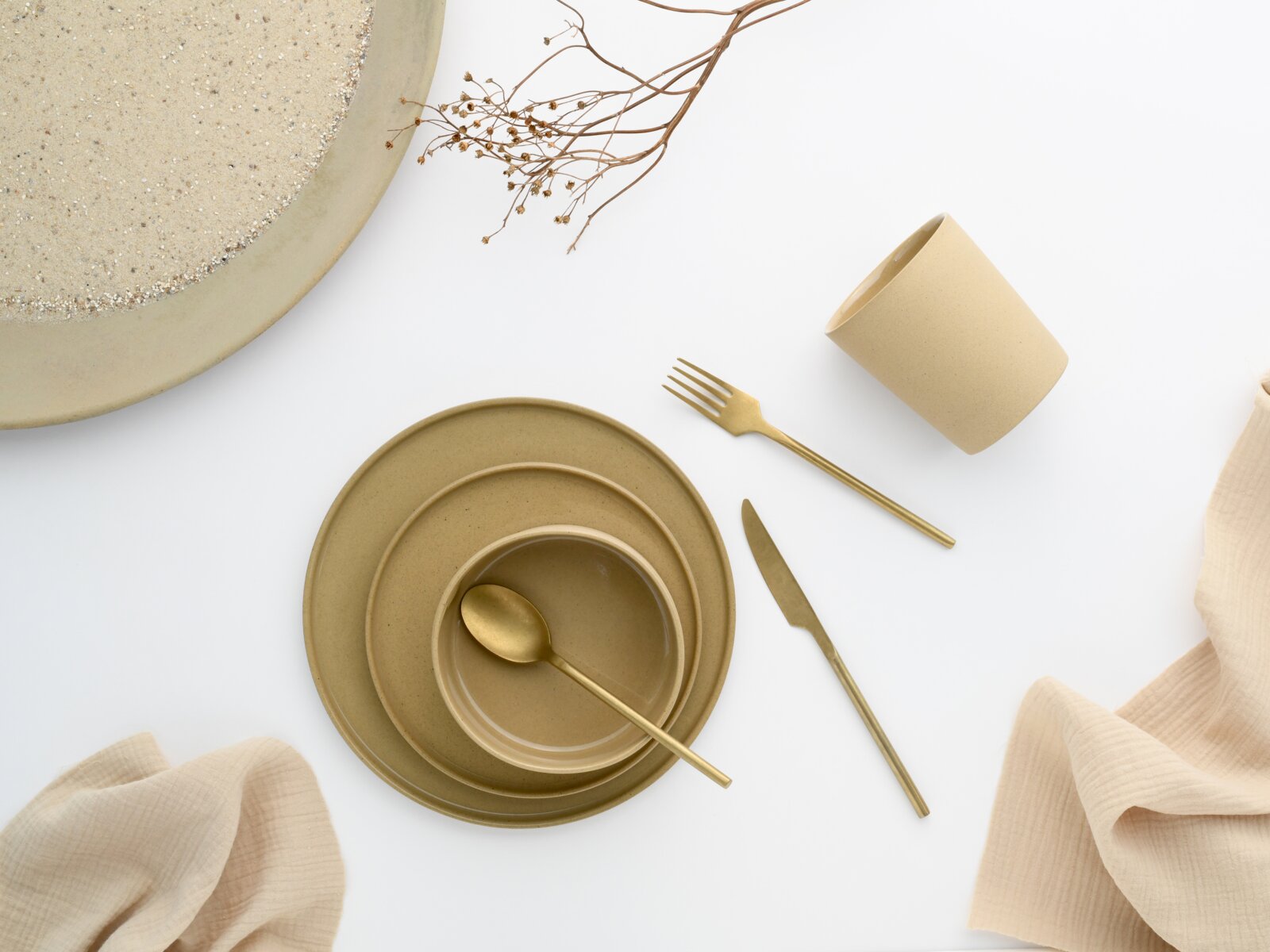 Minimalistisches beiges Keramik-Geschirrset mit Tellerstapel, Schale und Becher sowie goldfarbenem Besteck auf weißem Hintergrund, Draufsicht (Flatlay)