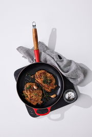 Bratpfanne 28 cm Kirschrot mit Holzgriff, von oben fotografiert, mit gebratenen Steaks, Knoblauch und Kräutern, auf einem Schneidebrett mit Salz und Pfeffer.