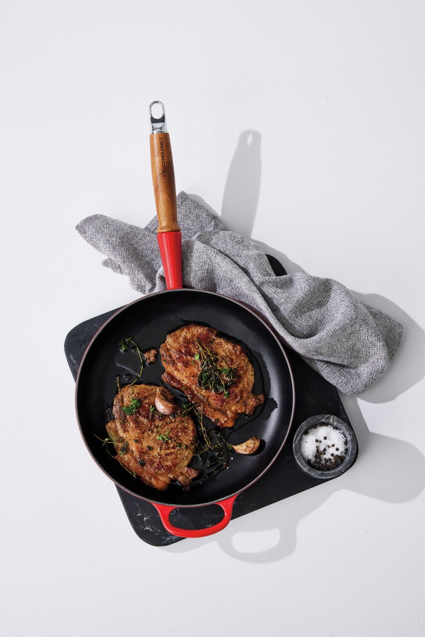 Koekenpan van 28 cm, kersenrood met houten handvat, gefotografeerd van bovenaf, met gebakken steaks, knoflook en kruiden, op een snijplank met zout en peper.