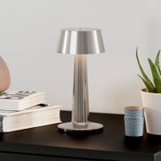 Moderne Tischleuchte aus gebürstetem Edelstahl mit konischem Schirm auf schwarzem Sideboard, seitliche Frontansicht leicht von oben, neben Büchern und Pflanze