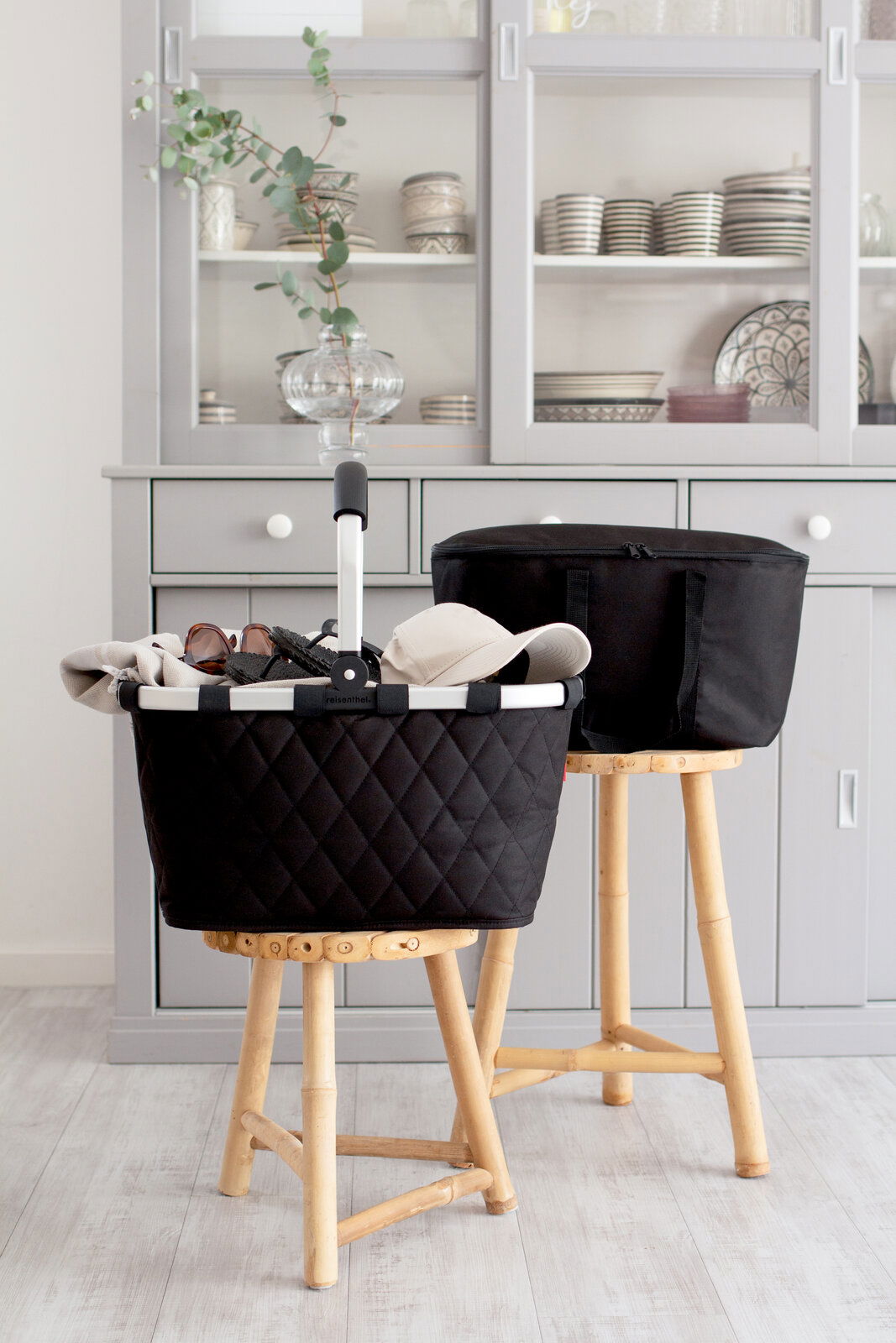 Schwarzer Rhombus-Einkaufskorb auf einem Hocker, gefüllt mit Accessoires, in einem modernen Raum mit grauem Schrank im Hintergrund, seitliche Perspektive.