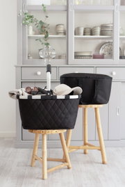 Schwarzer Rhombus-Einkaufskorb auf einem Hocker, gefüllt mit Accessoires, in einem modernen Raum mit grauem Schrank im Hintergrund, seitliche Perspektive.