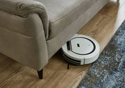 Nahaufnahme eines grauen Sofas mit einem Saugroboter, der darunter fährt, auf einem Holzboden mit Teppich.