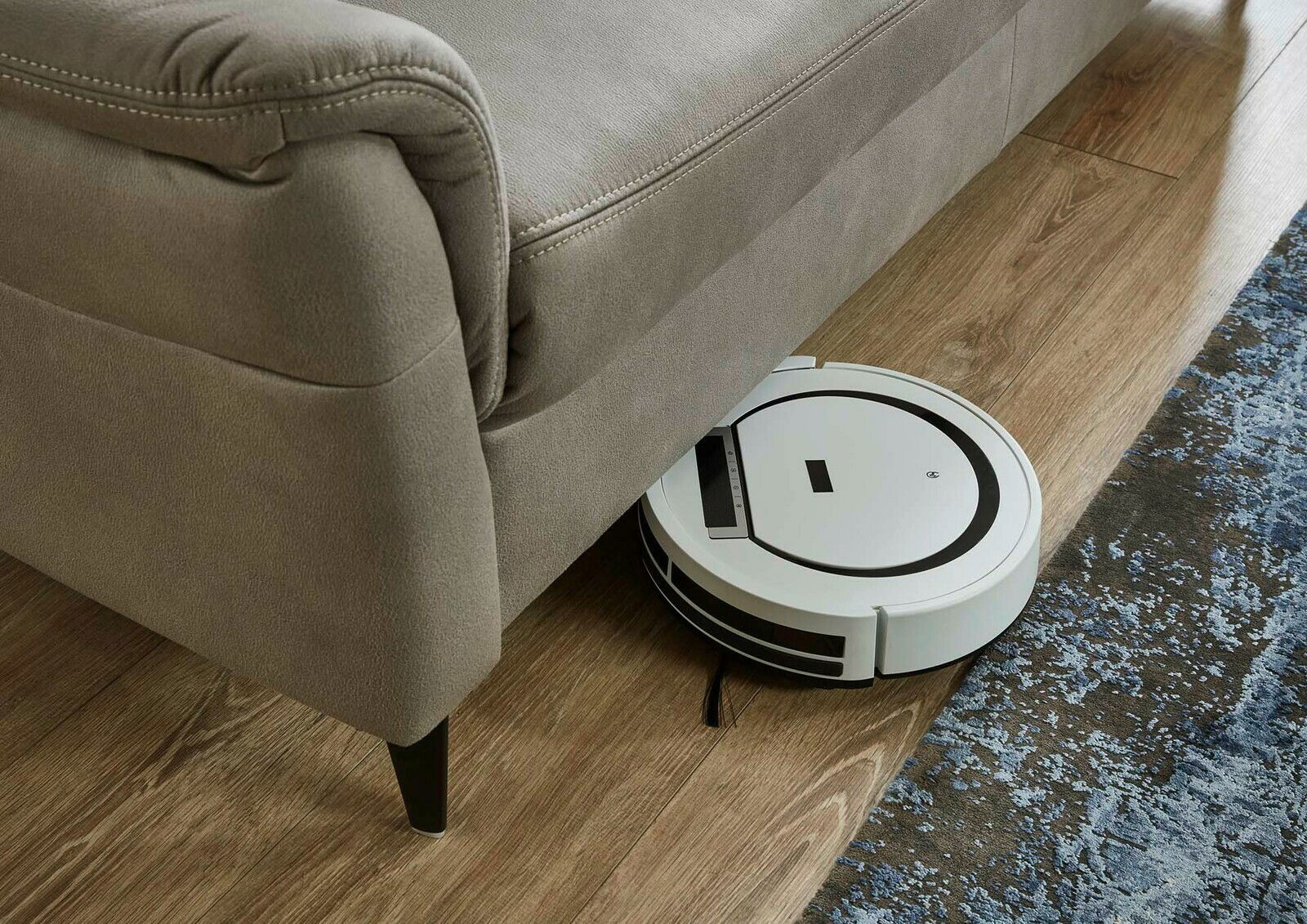 Nahaufnahme eines grauen Sofas mit einem Saugroboter, der darunter fährt, auf einem Holzboden mit Teppich.
