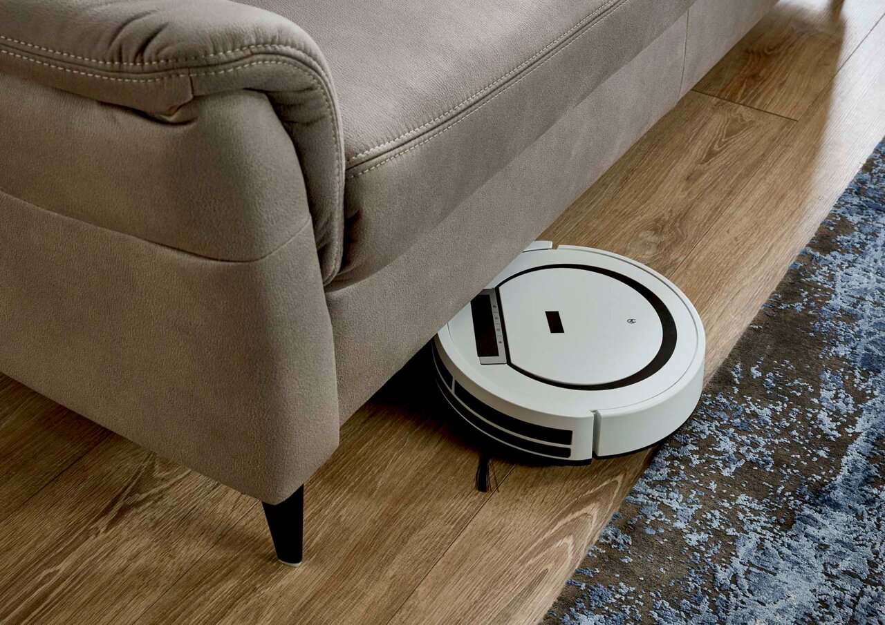 Close-up van een grijze bank met een robotstofzuiger die eronder rijdt, op een houten vloer met tapijt.