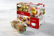 Zwei Muffins in WECK-Gläsern mit bunten Streuseln, vor einer Verpackung mit der Aufschrift 'Lecker genießen Muffin im Original WECK-Glas', auf einer hellen Holzoberfläche. Perspektive: leicht von oben.
