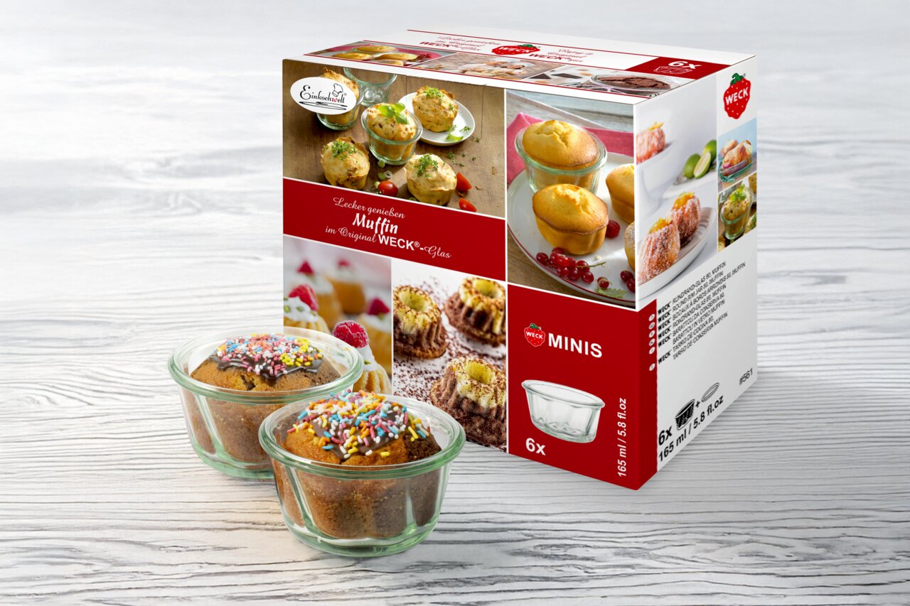 Zwei Muffins in WECK-Gläsern mit bunten Streuseln, vor einer Verpackung mit der Aufschrift 'Lecker genießen Muffin im Original WECK-Glas', auf einer hellen Holzoberfläche. Perspektive: leicht von oben.