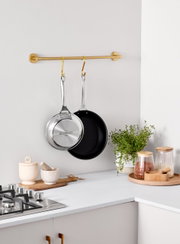 Twee 3-PLY-pannen, één met een roestvrijstalen bodem en één met een zwarte antiaanbaklaag, hangen aan een gouden stang in een moderne keuken. Zijaanzicht.