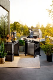 Schwarzer Gasgrill mit geöffnetem Deckel und Rauch auf einer sonnigen Terrasse, seitliche Ablage mit Grillgut; Perspektive: frontale leicht seitliche Ansicht auf Augenhöhe.