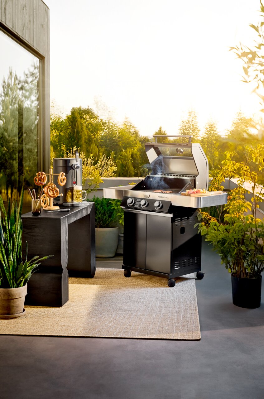 Rösle BBQ-Station VIDERO G3 inkl. Abdeckhaube Schwarzer Gasgrill mit geöffnetem Deckel und Rauch auf einer sonnigen Terrasse, seitliche Ablage mit Grillgut; Perspektive: frontale leicht seitliche Ansicht auf Augenhöhe.