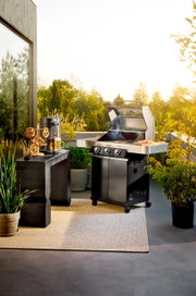 Zwarte gasbarbecue met open deksel en rook op een zonnig terras, met aan de zijkant een legplank met barbecuevoedsel; perspectief: vooraanzicht, licht van opzij, op ooghoogte.