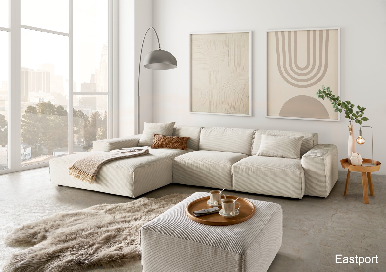 Lounge Handmade Hoekbank LH-EDINA Moderne, helle Wohnzimmerlandschaft mit einer großen, beigen Eckcouch und einem passenden Hocker, dekoriert mit Kissen und Decken. Im Vordergrund ein flauschiger Teppich, im Hintergrund große Fenster mit Stadtblick. Perspektive: Frontalansicht.