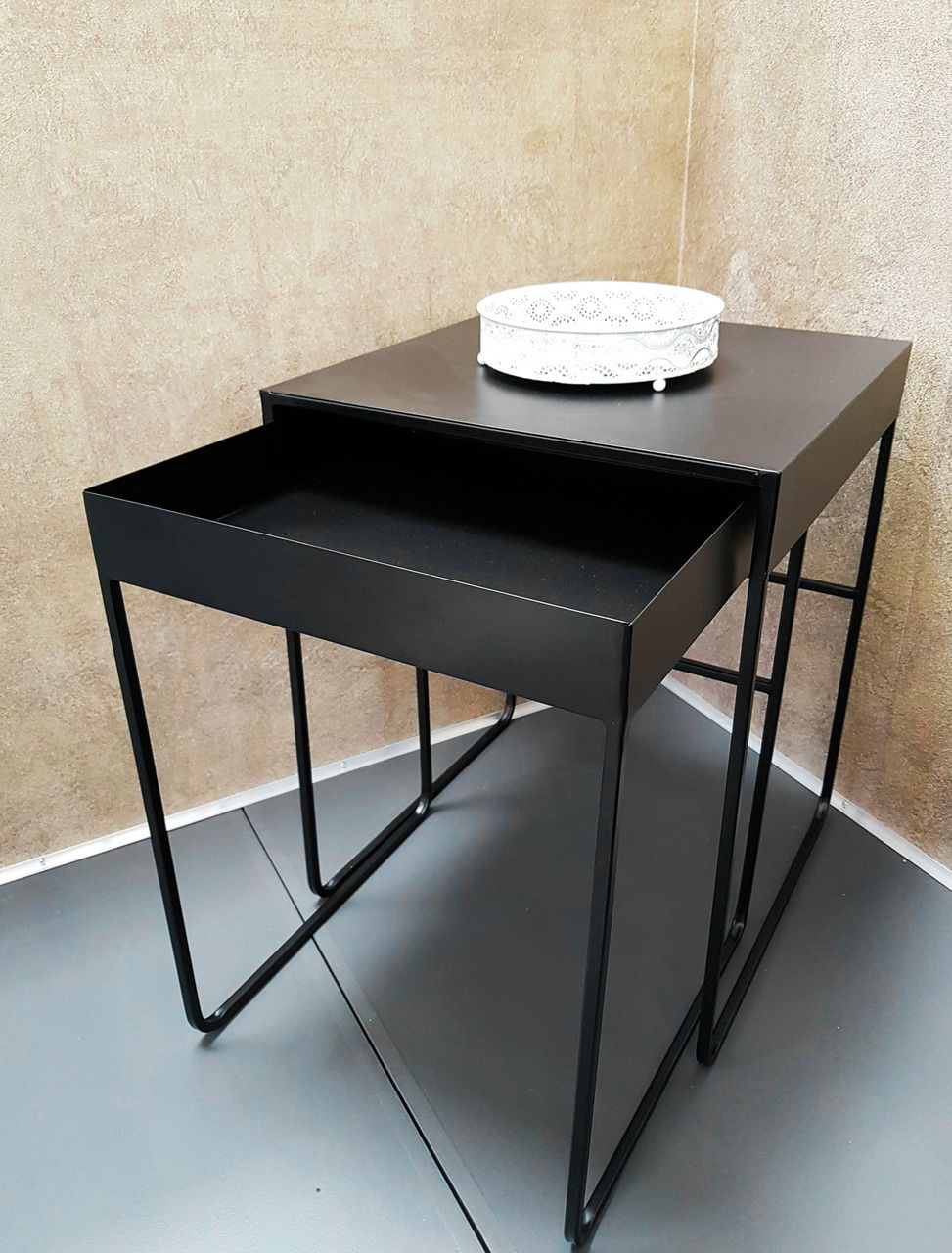 Zweier-Set Beistelltische in schwarzem Metall, übereinander gestapelt, mit einem dekorativen weißen Tablett auf dem oberen Tisch, seitliche Perspektive.