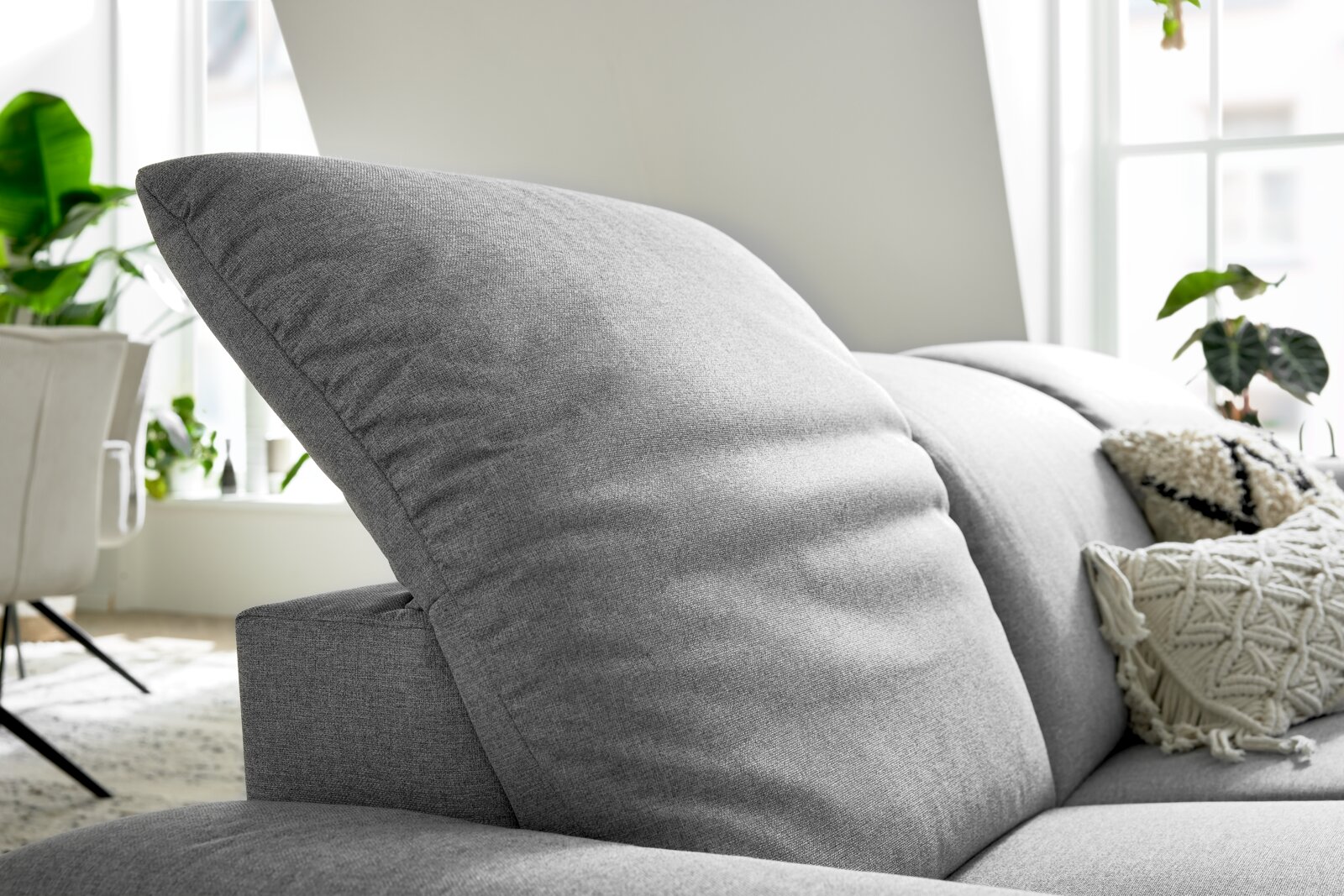 Nahaufnahme eines grauen Longchairs mit weichen Kissen, im Hintergrund sind Pflanzen und ein Fenster zu sehen.