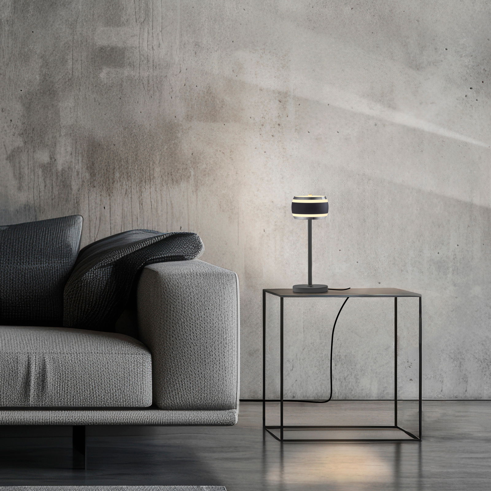 Moderne Akku-Tischleuchte auf einem minimalistischen Beistelltisch neben einem grauen Sofa, seitliche Perspektive.