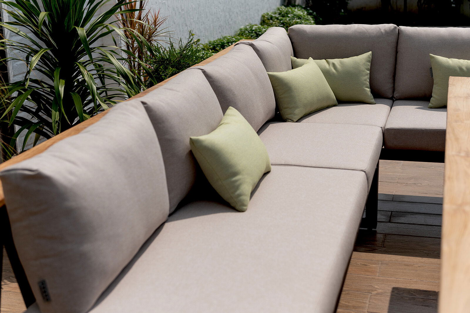 Eck-Sitzbank mit beigen Polstern und grünen Kissen, umgeben von Pflanzen, aus seitlicher Perspektive fotografiert.