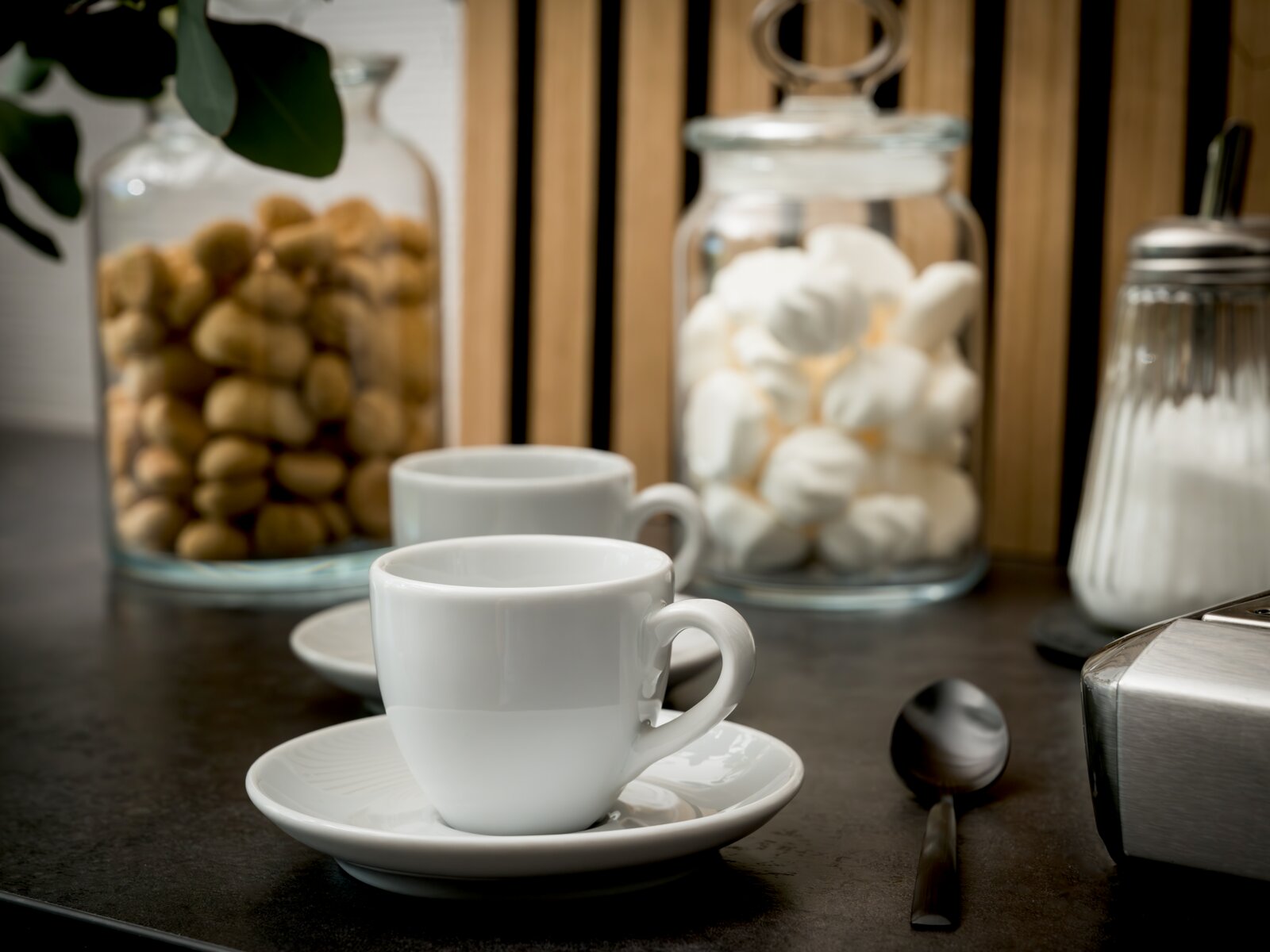 Zwei weiße Espressotassen mit Untertassen auf dunkler Arbeitsplatte, daneben Löffel und Zuckerstreuer; im Hintergrund Glasbehälter mit Keksen und Marshmallows, Perspektive: leicht von vorne und auf Augenhöhe