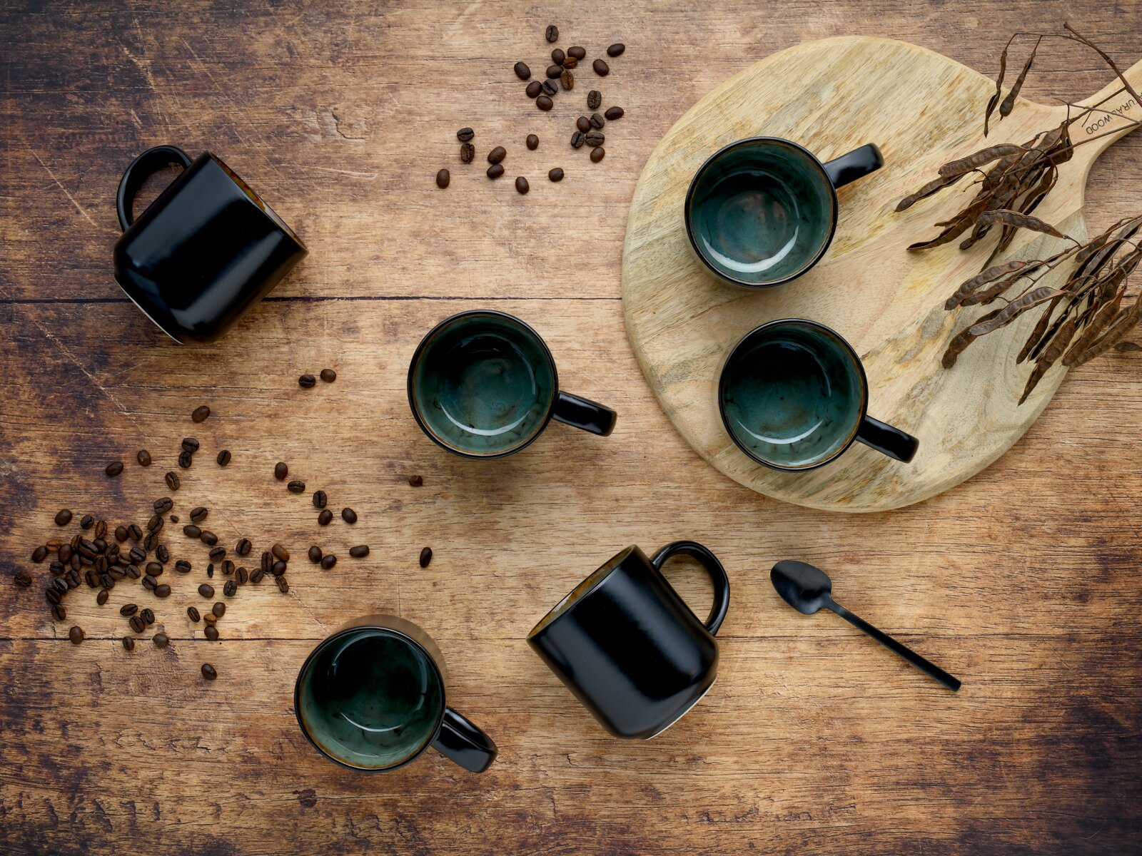 Draufsicht auf schwarze Keramik-Kaffeetassen mit grün glasiertem Innenraum auf rustikalem Holztisch, mit Kaffeebohnen, Holzbrett und Löffel dekorativ arrangiert.