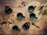 Draufsicht auf schwarze Keramik-Kaffeetassen mit grün glasiertem Innenraum auf rustikalem Holztisch, mit Kaffeebohnen, Holzbrett und Löffel dekorativ arrangiert.