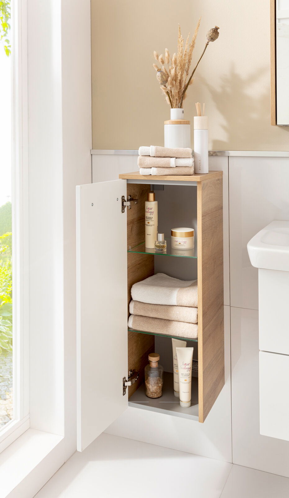Moderner Midischrank in Holzoptik mit geöffneter Tür, gefüllt mit Handtüchern und Pflegeprodukten, seitliche Perspektive