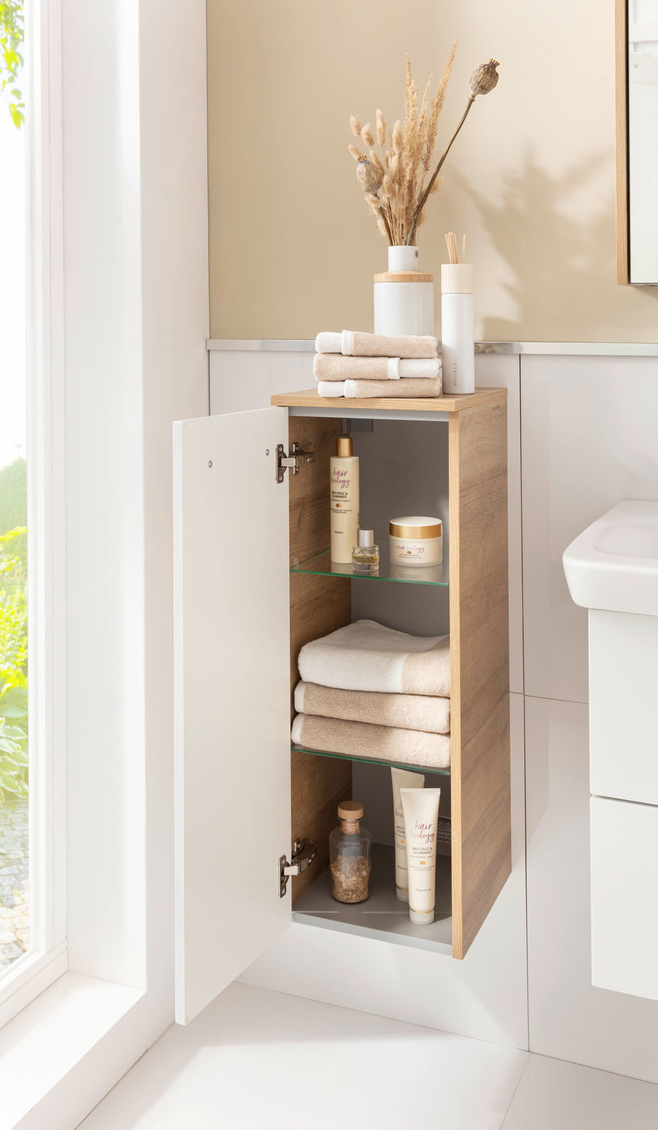 Moderner Midischrank in Holzoptik mit geöffneter Tür, gefüllt mit Handtüchern und Pflegeprodukten, seitliche Perspektive