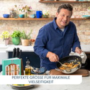Jamie Oliver in einer Küche, hält eine Servierpfanne mit Essen in der Hand, seitliche Perspektive.