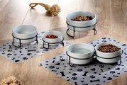 Napfset mit zwei weißen Schalen auf schwarzen Metallständern, gefüllt mit Wasser und Trockenfutter, auf einer Matte mit Pfoten- und Knochenmotiven, aus seitlicher Perspektive fotografiert.