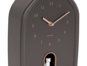Moderne Wanduhr in dunklem Gehäuse mit goldenen Zeigern und Zahlen, seitliche Perspektive, mit einer kleinen weißen Katze in einer Öffnung unten.