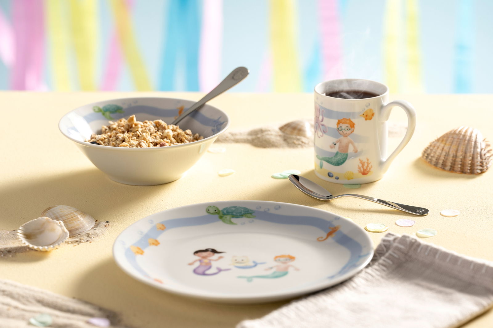 Kindergeschirrset mit Meeresmotiven, bestehend aus einem Teller, einer Schale und einer Tasse, auf einem Tisch mit Muscheln und buntem Hintergrund, seitliche Perspektive.