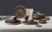 Ein stilvoll arrangiertes Set aus Keramiktellern und Schalen in Erdtönen, mit einem großen Platzteller im Hintergrund. Perspektive: Frontalansicht auf das Geschirrset auf einem Tisch.