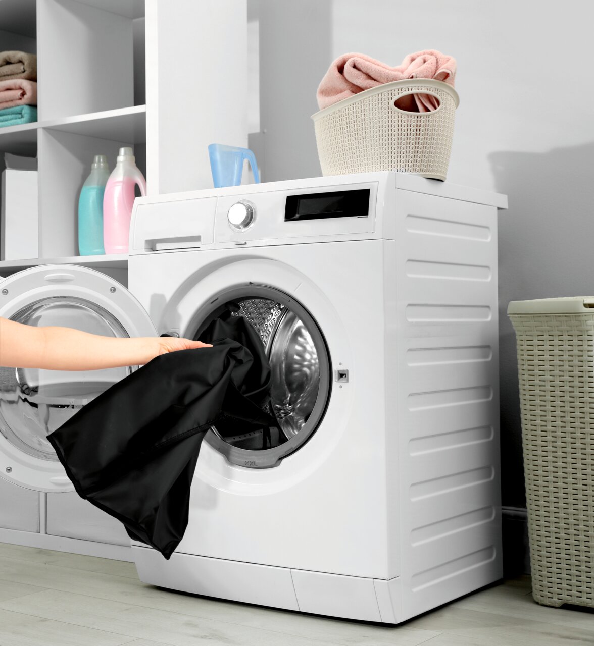 Frontansicht einer modernen Waschmaschine mit geöffneter Tür, in die eine Person schwarze Wäsche legt. Oben auf der Maschine steht ein Korb mit gefalteten Handtüchern.