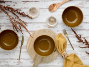 Drei olivgrüne Keramikschalen mit goldfarbenem Rand auf weißer Holztafel, mit Holzbrett, Besteck, Holzlöffel, gelber Serviette und Deko-Zweigen, Draufsicht (Flatlay)