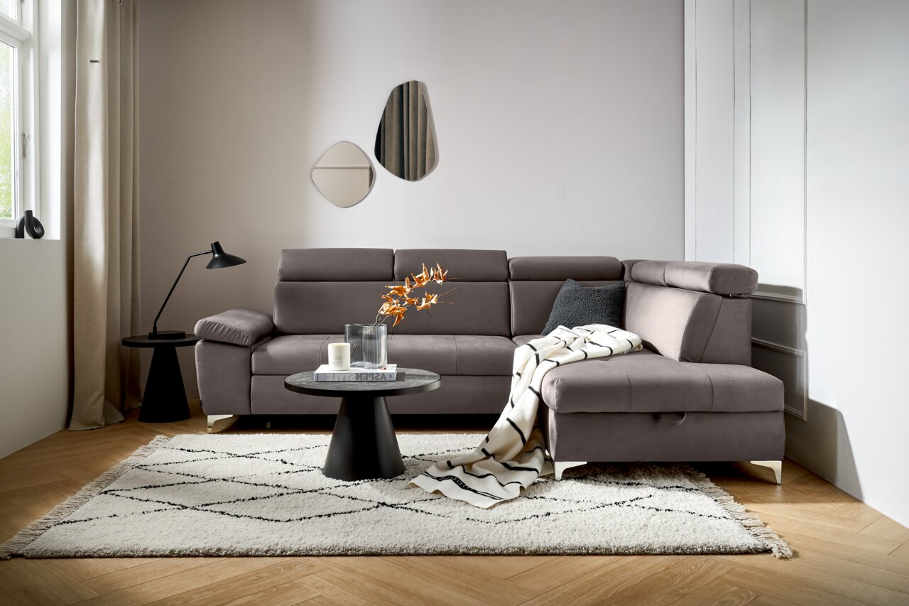 Een moderne grijze hoekbank in een stijlvol ingerichte woonkamer, gezien vanaf de voorkant. De bank is versierd met een kussen en een deken, en staat te midden van een vloerkleed, een salontafel en een staande lamp.