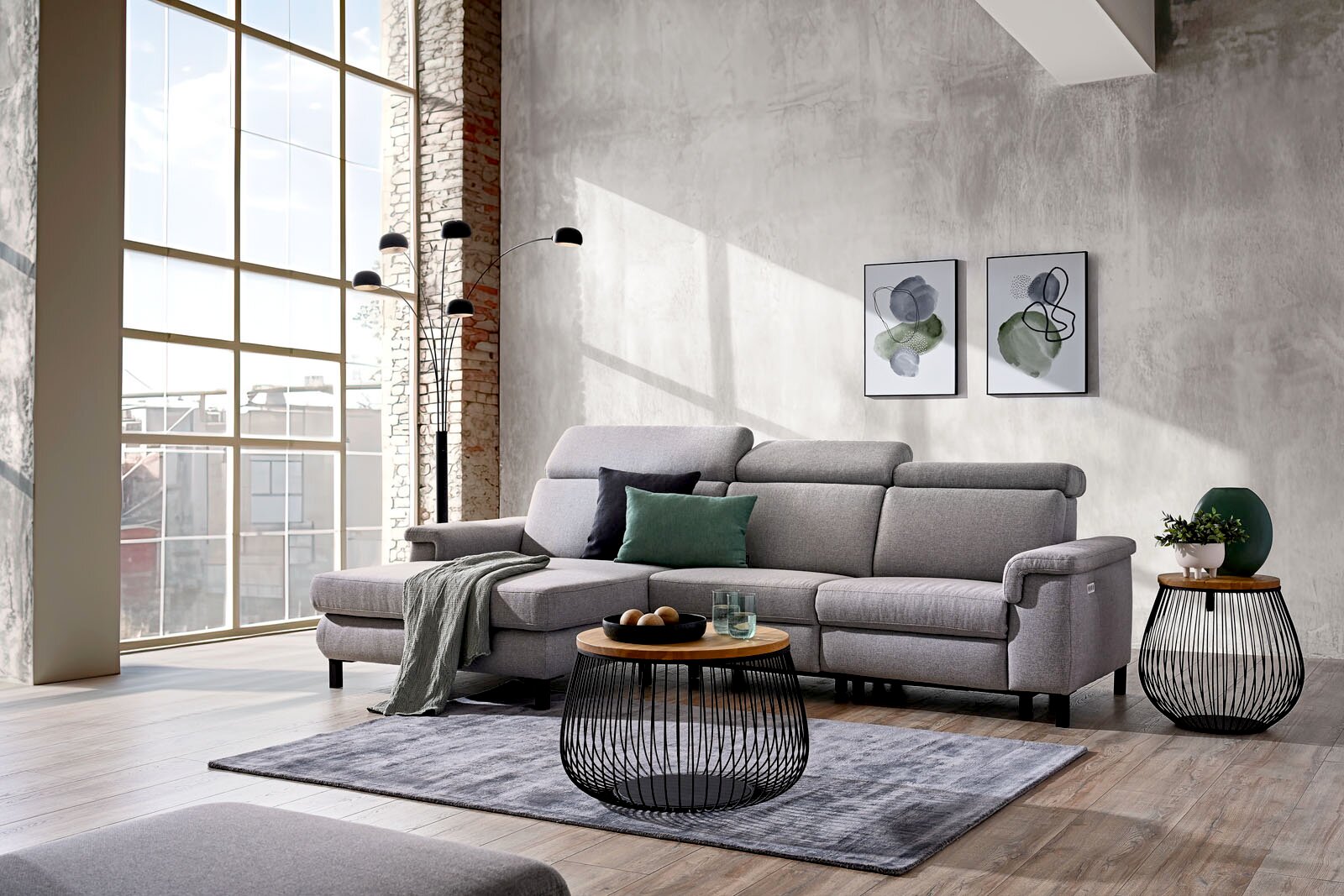 Graues Ecksofa mit Ottomane, verstellbaren Kopfstützen und dekorativen Kissen, aus frontaler Perspektive in einem modernen, lichtdurchfluteten Loft-Wohnzimmer gezeigt.