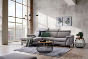 Graues Ecksofa mit Ottomane, verstellbaren Kopfstützen und dekorativen Kissen, aus frontaler Perspektive in einem modernen, lichtdurchfluteten Loft-Wohnzimmer gezeigt.
