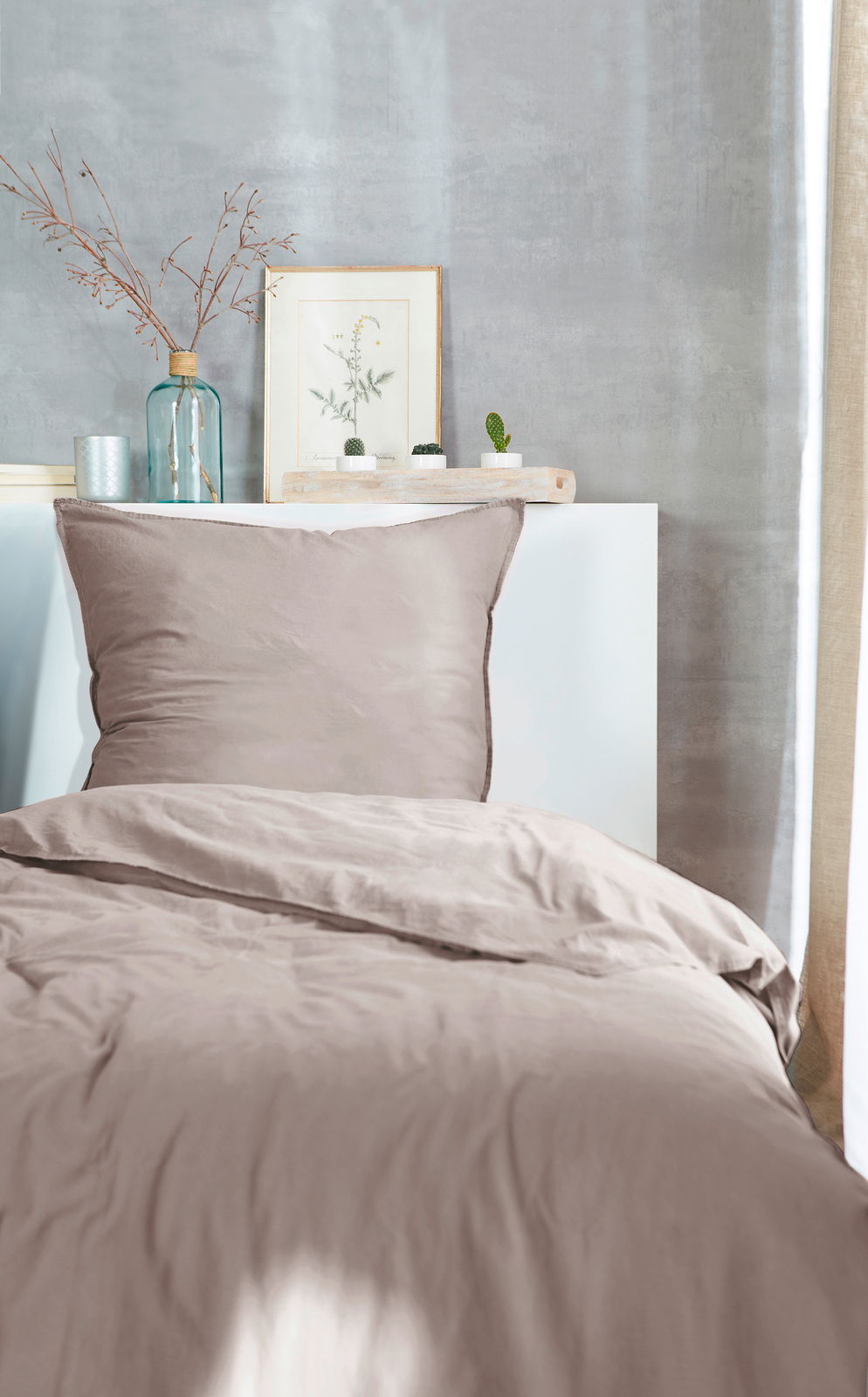 Bettwäsche-Set in sanftem Beige, bestehend aus einem Kissenbezug und einem Bettdeckenbezug, auf einem Bett mit dekorativem Hintergrund aus Pflanzen und Vasen, aus der Vorderansicht fotografiert.