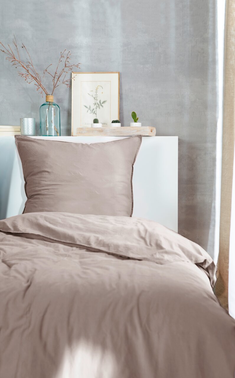 Bettwäsche-Set in sanftem Beige, bestehend aus einem Kissenbezug und einem Bettdeckenbezug, auf einem Bett mit dekorativem Hintergrund aus Pflanzen und Vasen, aus der Vorderansicht fotografiert.