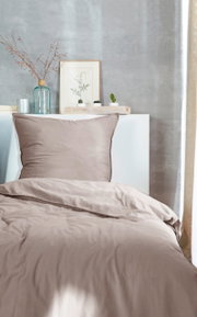 Bettwäsche-Set in sanftem Beige, bestehend aus einem Kissenbezug und einem Bettdeckenbezug, auf einem Bett mit dekorativem Hintergrund aus Pflanzen und Vasen, aus der Vorderansicht fotografiert.