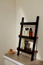 Schuin gefotografeerde, zwarte laddervormige badkamerplank met houten legplanken op een wastafelblad in een hoek, met parfumflessen, handcrème, een tube en een horloge; op de voorgrond een houten borstel.