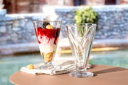 Zwei hohe Glasschalen, eine gefüllt mit Dessert, auf einem Tisch im Freien, seitliche Perspektive.