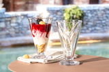 Zwei hohe Glasschalen, eine gefüllt mit Dessert, auf einem Tisch im Freien, seitliche Perspektive.