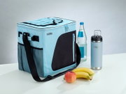 Lichtblauwe Thermos-koeltas met zwarte schouderriem en zijvak, vanuit schuin vooraanzicht gefotografeerd naast een drinkfles, waterfles, appel en bananen.