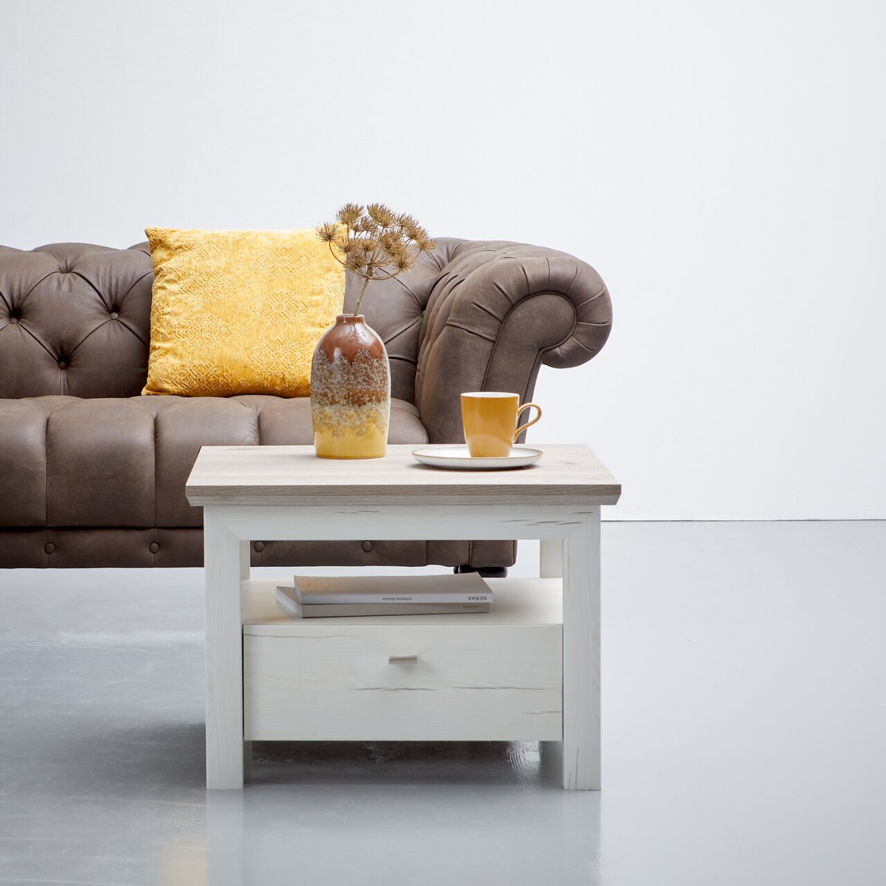 Bijzettafel CAMRON in lichte houtlook met lade, versierd met een vaas en een kopje, voor een bruine bank, vooraanzicht.