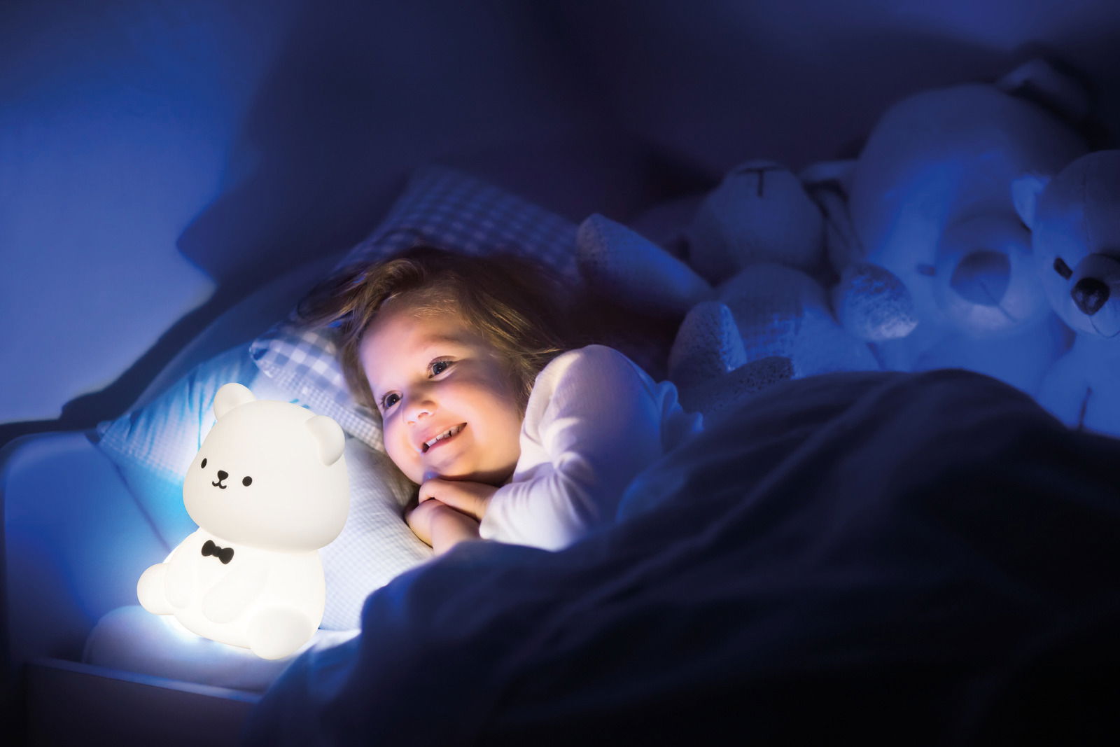 Ein Kind liegt im Bett und lächelt, während es ein leuchtendes Bären-Nachtlicht betrachtet. Das Nachtlicht hat die Form eines sitzenden Bären mit einer schwarzen Fliege. Die Szene ist in einem dunklen Raum mit sanfter blauer Beleuchtung aufgenommen.