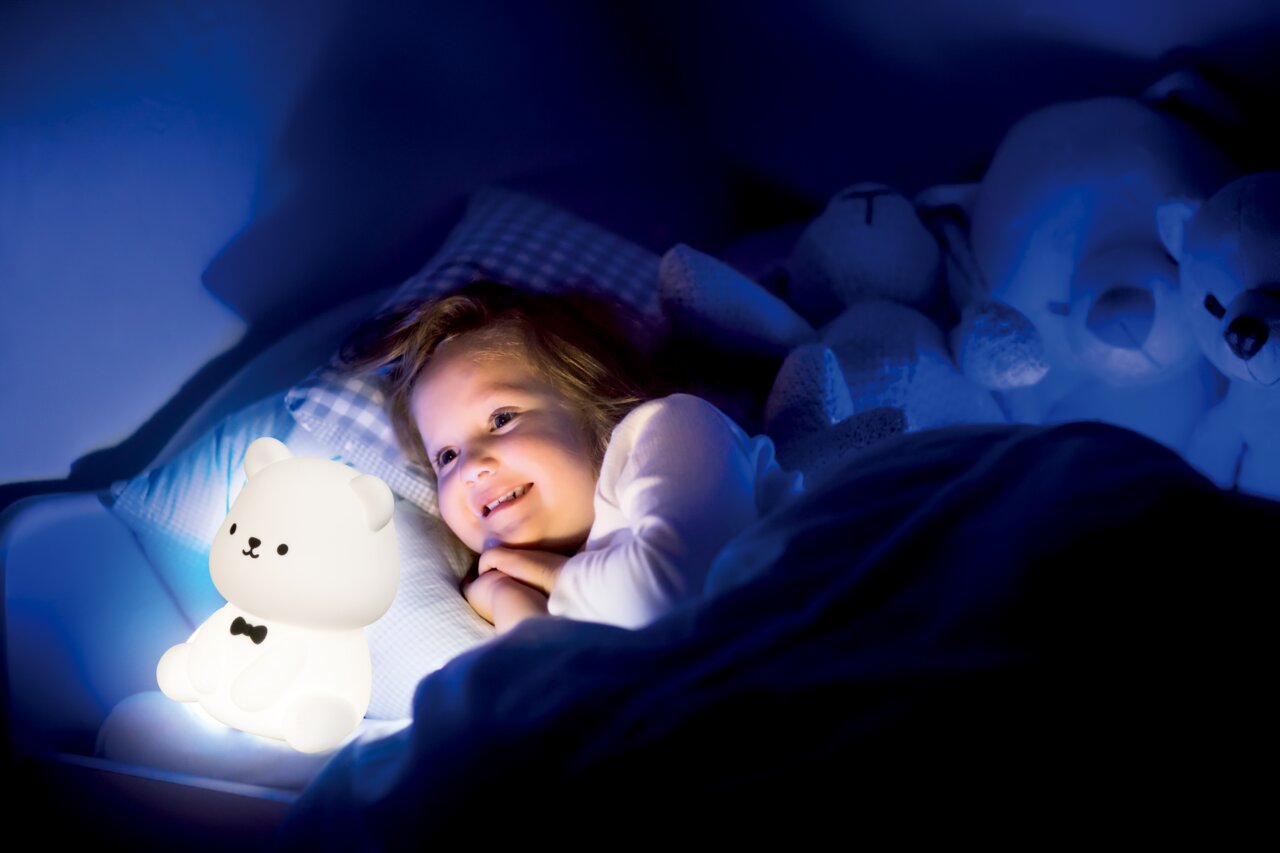 Ein Kind liegt im Bett und lächelt, während es ein leuchtendes Bären-Nachtlicht betrachtet. Das Nachtlicht hat die Form eines sitzenden Bären mit einer schwarzen Fliege. Die Szene ist in einem dunklen Raum mit sanfter blauer Beleuchtung aufgenommen.