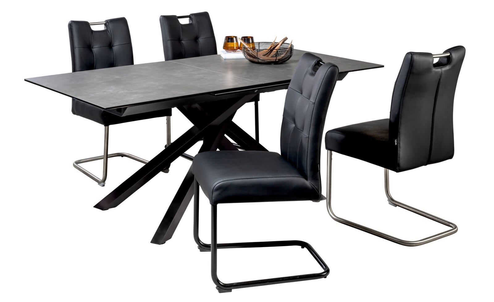 Modernes Esszimmer-Set mit ausziehbarem Tisch in Betonoptik und schwarzem X-Gestell sowie vier schwarzen Polsterstühlen mit Schwinggestell, fotografiert in schräger Vorderansicht vor weißem Hintergrund