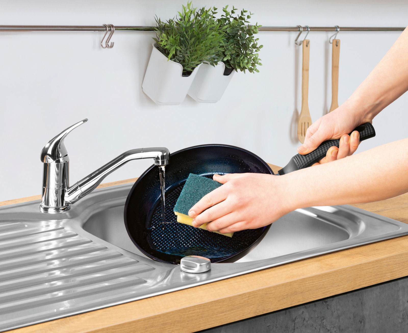 Eine Person reinigt eine Pfanne unter fließendem Wasser in einer Küchenspüle. Die Perspektive zeigt die Pfanne von oben, während sie mit einem Schwamm geschrubbt wird.
